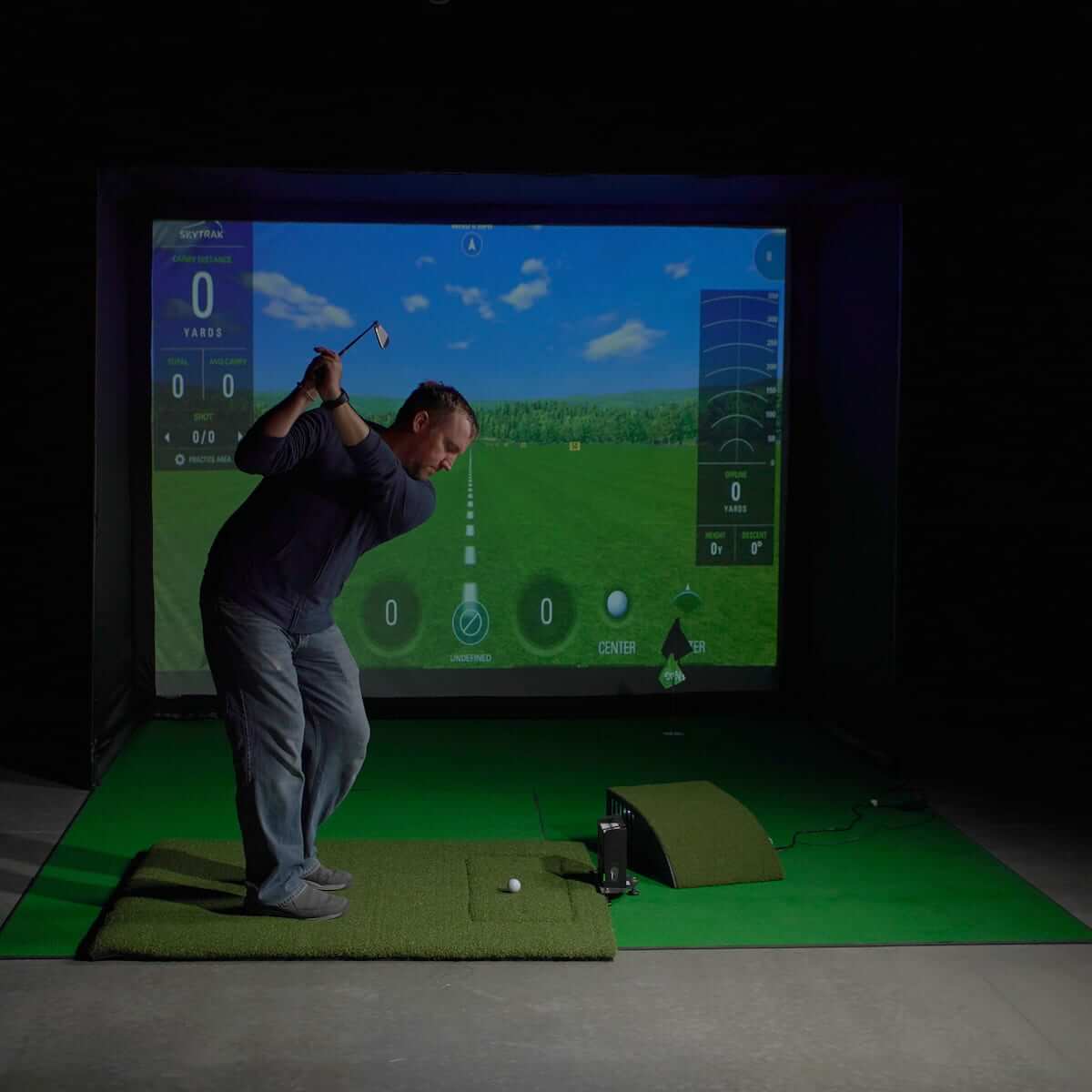 Golfer using Carl's Place HotShot Golf Hitting Mat with golf simulator setup in a home environment.